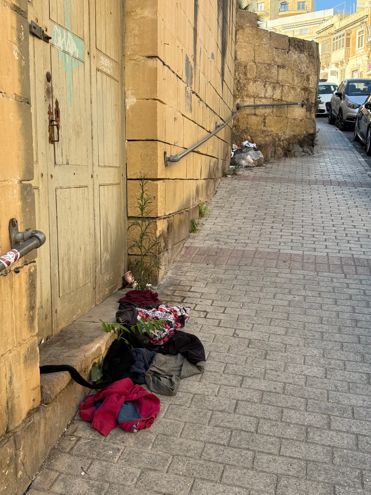 A weathered Sliema street: cracked pavement, dark sandbags lining the gutter, peeling buildings