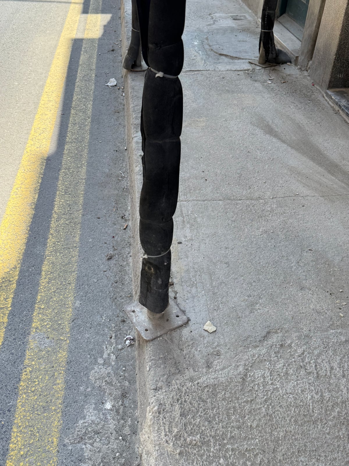 Gallery Three: a black pole whose base plate balances on the edge of the kerb, anchored to nothing