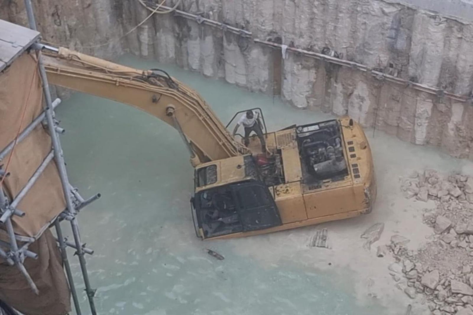 A worker stands on a partially submerged excavator in a flooded foundation pit